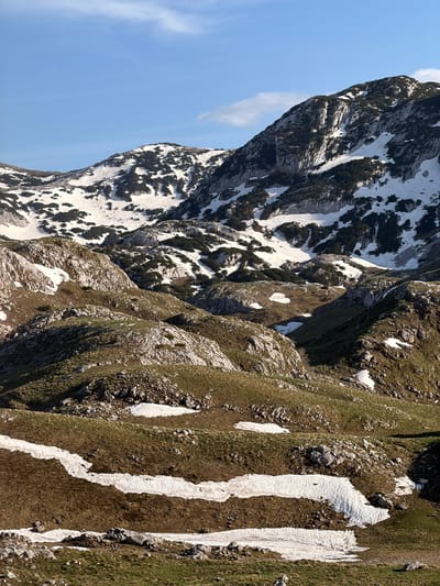 The image depicts a mountain landscape in Boričje, Montenegro. The left side of the image shows a rocky mountain peak, partially covered in snow, under a blue sky with a wisp of cloud. To the right of the peak, there are steep, grassy slopes with patches of snow, leading down towards the foreground. Rocks are scattered throughout the slopes. The image has a strong diagonal composition.