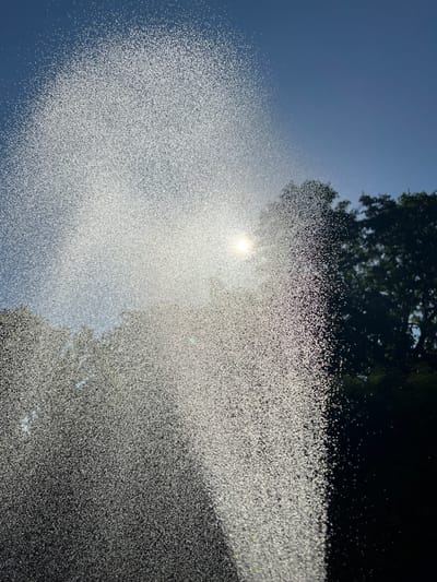 The image shows a close-up view of water spraying from a sprinkler against a bright blue sky. The sun is visible through the spray, creating a shimmering, iridescent effect with numerous tiny water droplets catching the light. The water spray forms a dense curtain in the foreground, with individual droplets clearly distinguishable, especially where the sunlight hits them. In the background, dark silhouettes of trees are visible against the sky, suggesting an outdoor setting, possibly a garden or park. The time of day appears to be midday or late afternoon, given the bright sunlight and the angle of the sun. The overall impression is one of a refreshing, bright, and dynamic scene, likely captured during warm weather. There is no visible text in the image.