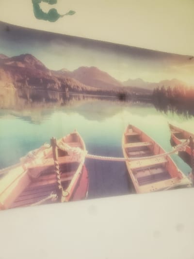 The image displays a photograph or print depicting a serene landscape, with the bottom third of the frame obscured by a bright white, featureless foreground, possibly a surface on which the image rests or is reflected. The print itself occupies the upper two-thirds of the visible area.
**Subjects and Objects (within the print):** The central subjects are three wooden rowboats or canoes floating on a calm body of water, likely a lake. Two boats are prominent in the foreground, parallel to each other, and are connected by a thick, light-colored rope. A third boat is partially visible on the far right. The water's surface reflects the sky and distant landforms. In the background, a range of mountains, covered with trees, rises under a sky exhibiting warm hues, suggesting either a sunrise or sunset.
**Actions and Interactions (within the print):** The boats appear stationary, tied together. The water reflects the environmental elements above it.
**Environment (of the photograph itself):** The entire image is captured at a slight downward angle. The print displays a muted, possibly vintage, color palette. Above the print, an irregular dark green mark is visible on a light, indeterminate background. A faint diagonal dark line crosses the upper portion of the print, possibly a shadow or structural element from the immediate surroundings. The bright white foreground shows slight irregularities, possibly dust or minor surface imperfections.
**Location:** While the depicted landscape is a generic mountainous lake scene not specifically identifiable with the geography of Saint John, Canada, this image is presented within the context of being physically located or captured within the city of Saint John, Canada. The specific address or precise location within Saint John where this photograph or print is situated is not discernible from the image content.