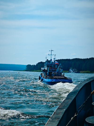 A blue tugboat, identified by the Russian text "ПЛАТОBOД 717" on its stern, is seen from the perspective of another vessel in the foreground. The tugboat is creating a significant wake as it moves through choppy, dark blue water. In the distance, a dense, dark green forest lines the far bank of a wide river, meeting a hazy, blue sky. Two buoys are visible in the water between the two vessels. Another, smaller white vessel is further in the distance, behind the tugboat. The overall scene suggests a sunny day on a Russian river.
