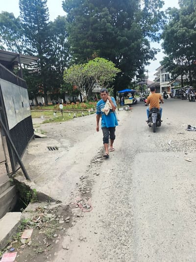A man in a blue shirt and dark pants walks along the edge of a road, carrying a white cloth bag over his shoulder. The road has an uneven edge, with a drainage ditch and metal grate to the left. A person on a scooter drives away on the road, and there are parked scooters in the distance. Trees and buildings line the background, and a small food cart is visible next to the walking man.