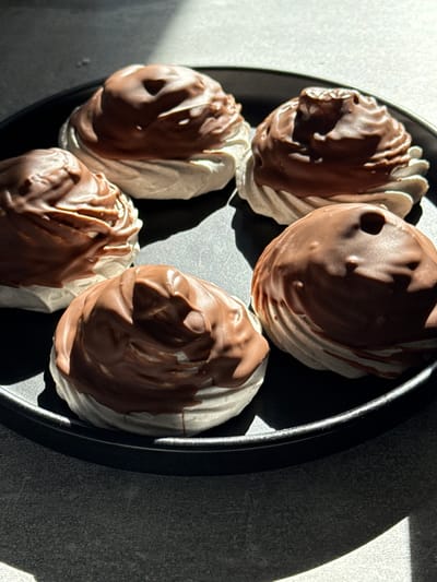 The image presents five individual confectionery items arranged on a dark, circular plate. Each item consists of a light-colored, possibly meringue or marshmallow-like, base with a textured, piped surface, almost entirely covered by a thick, smooth layer of milk chocolate. The chocolate coating shows slight irregularities and drips where it has solidified.
The plate is positioned on a gray, textured surface, appearing to be a countertop or table with a granular or speckled finish. Strong, directional lighting, likely natural, originates from the upper left of the frame, casting prominent, elongated shadows of the desserts and the plate onto the surface below. The background directly above the plate is a blurred, lighter gray tone. No people, actions, or interactions are depicted.
No specific geographical location, such as the city of Бордуки in Russia, can be determined from the visual information present in the image.