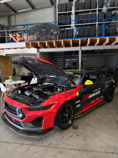 The scene depicts an interior view of a commercial garage or workshop located in Meuspath, Germany. The primary subject is a highly modified racing car, resembling a Ford Mustang, parked on a grey concrete floor. The vehicle's livery is predominantly red on the front section, transitioning to black along the sides, with bright yellow accents on the side mirrors, brake calipers, and body trim lines. Sponsor logos such as "NANKANG MOTORSPORT," "VULCAN ALPHA," and "RTR VEHICLES" are visible on its exterior. The car's hood is open, exposing a performance-oriented engine bay with a prominent air intake system and various mechanical components. The front features an aggressive splitter with a yellow edge.
In the background, on the ground level to the left, a small break area includes a white round table, two white chairs, a green plastic bottle, and a clear glass. A sink and a checkered towel are visible beyond the table.
Above the main floor, a mezzanine level with a blue metal railing extends across the upper portion of the facility. This mezzanine functions as a storage area, featuring metal shelving units densely packed with stacks of black tires. A dark, possibly carbon fiber, vehicle body panel rests on the mezzanine floor near the railing. Metal stairs leading to the mezzanine are partially visible on the right side of the frame. The facility is illuminated by overhead lighting. No individuals are present in the image.