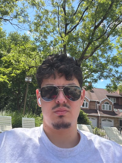 A close-up, portrait-style photograph captures a person outdoors on a bright, sunny day. The individual, seen from the chest up, has dark, curly hair and is wearing reflective aviator-style sunglasses. A mustache and goatee are visible on their face. White wireless earbuds are visible in both ears. They are wearing a plain white t-shirt.
The background reveals a lush green environment with numerous trees whose leaves are brightly lit by the sun, indicating clear blue skies overhead. Beyond the foliage, a multi-story building or house is partially visible. This structure features brown siding, multiple windows with white frames, and distinct architectural details, including a prominent curved window. Below the trees and to the side, several white slatted outdoor chairs, possibly Adirondack style, are arranged on what appears to be a patio or lawn. The overall scene suggests a relaxed, casual setting in a residential or resort-like area. There is no visible text in the image.