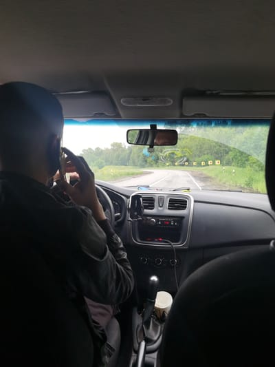 The image captures the interior of a vehicle from a rear passenger's perspective, looking forward through the windshield onto a rural road in Кварса, Russia.

A male driver with short, dark hair is visible from the back of his head and right shoulder in the left foreground. His right hand holds a silver-colored smartphone to his right ear, indicating he is engaged in a phone conversation. His left hand rests on the black steering wheel. He is wearing a dark, long-sleeved garment, possibly a jacket.

The car's interior features a dark grey dashboard and a manual transmission gear stick with an illuminated white top, positioned between the driver and the front passenger seat. A white disposable cup with a lid sits in a cup holder near the gear stick. The center console houses a car radio displaying red text, and a red light is visible near a connected cable. A black phone mount is affixed to the dashboard to the right of the steering wheel. The rearview mirror, centrally mounted, displays a blurred reflection, and a decorative, intricate knot-like charm hangs from it. Above the windshield, grey sun visors and a grey textured roof liner are visible. The upper right portion of a dark-colored front passenger seat is partially visible in the right foreground.

Beyond the windshield, a two-lane asphalt road curves to the left. The road has a solid white line on its left edge and a dashed white line separating the lanes. Along the right side of the road, a series of yellow square road signs with black left-pointing arrows indicate a sharp bend ahead, accompanied by white marker posts. The environment is characterized by dense green trees and foliage lining both sides of the road, with some yellow wildflowers visible in the foreground greenery. The sky is overcast.