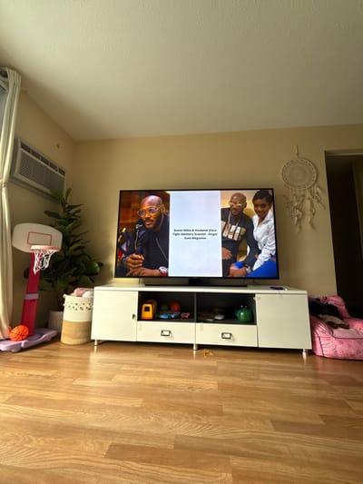 This media file captures an indoor living space, likely a living room or family room. The main focal point is a large flat-screen television displaying what appears to be a news report or entertainment segment. To the right of the TV, there's a white media console with open shelves and closed compartments, holding various items including children's toys like small basketballs and a yellow toy truck. A decorative white woven dream catcher hangs on the wall above the TV.
On the left side of the room, a large green leafy plant in a pot stands on the floor. Adjacent to it, a small pink and white toy basketball hoop with a miniature basketball is set up, suggesting the presence of children. A window air conditioning unit is visible on the far left wall. In the background, to the right of the media console, a pink bean bag chair or pet bed is present. The room features light-colored walls and light-toned wood-look flooring.
The television screen shows images of people; the upper half features two men, one in a white shirt and another in a dark shirt with a chain, while the lower half displays a man with glasses in a dark shirt. Overlaying the image on the screen is white text that reads: "Annie Idibia & Husband 2face", "Fight Adultery Scandal, Singer", and "Sues Magazine". The ambient light suggests it is daytime. The overall scene conveys a lived-in, casual domestic environment in Charlton, United States.