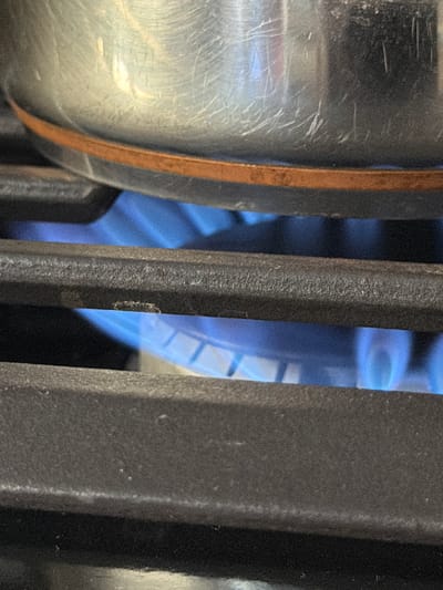 A close-up, eye-level shot captures a domestic scene focused on a gas stovetop. In the foreground on the left, a portion of a silver-colored metal pot or kettle is visible, showing numerous scratches, scuffs, and reflections on its surface, indicating wear. A rust-colored band or handle attachment runs vertically along its right side.
Dominating the center and right side of the frame are dark gray or black stove grate bars, likely made of cast iron, with a visibly rough, textured surface. These bars run vertically, creating narrow openings between them.
Through these gaps in the grate, a vibrant blue gas flame is clearly visible, suggesting an active burner heating the pot. The flame exhibits a circular pattern within its glow, hinting at the design of the burner head beneath. On one of the central grate bars, a small, light-colored mark or speck of debris can be seen.
The image does not show any people, visible text, or specific location cues beyond a kitchen setting. The lighting is artificial, suggesting an indoor environment.
