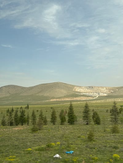 The image shows a landscape with a sky filled with clouds covering the left side of the image.
To the right of the sky and clouds is a mountain range. On the far side of the mountain range is what appears to be a quarry. A strip of trees borders the base of the mountain on the right.
On the right of the image is a field of grass. Litter, a blue plastic bag, and a white bag are visible in the grass.
The location is Karatay, Turkey.