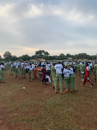 The image displays a large outdoor gathering in Kubi A, Nigeria, under an overcast sky. A substantial number of individuals, primarily young adults, are present on reddish-brown ground with sparse green grass. Most individuals wear a uniform consisting of a white t-shirt with "NYSC" branding in green, light green trousers, and bright orange-yellow shoes, identifying them as National Youth Service Corps members. They are standing and conversing in various groupings across the scene. In the mid-ground, two mobile vendor carts are visible; the left cart displays packaged snacks and drinks, while the right cart holds less identifiable goods. A child in an orange top and an adult in a reddish-brown garment stand near these carts. The background features multiple trees, a small, light-colored building, and utility poles. Scattered litter is visible on the ground.
