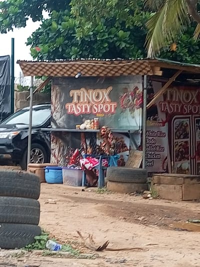 The image depicts a street scene in Oshogbo, Nigeria, featuring a food stall named "TINOX TASTY SPOT". The stall offers items such as White Rice, Dodo, Jollof Rice, Chicken, and Beef. A black vehicle is parked next to the stall. There are containers, food items, and personal belongings visible on and around the stall. In the foreground are stacks of tires and litter on a dirt ground. The background includes trees and foliage.