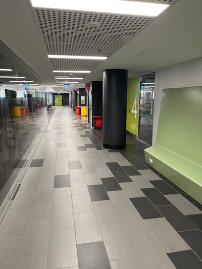 A long, brightly lit, modern interior corridor or hallway is depicted, viewed from a slightly elevated perspective. The image is oriented vertically, but depicts a horizontal space. The left side of the corridor features a white wall with extensive perforated paneling and prominent linear LED light fixtures running its length. The right side wall is covered in a pattern of rectangular tiles in varying shades of light grey and dark grey. Above the tiled wall, a light green panel extends along the ceiling.

The floor is dark and highly reflective, mirroring the overhead lights, the colorful objects, and the green sign further down the hall. In the mid-ground, a large, black cylindrical pillar stands prominently. Behind it, a tall, vertical green sign is visible with a large white number "4" and "S409" printed below it. A row of colored waste bins—black, red, and yellow—are lined up against the right wall, near the green sign. Further down the corridor, another blue sign with a white "4" is visible. The corridor appears empty, suggesting a quiet time. The architecture is clean, geometric, and functional, typical of modern institutional or commercial buildings in Brisbane City, Australia.
