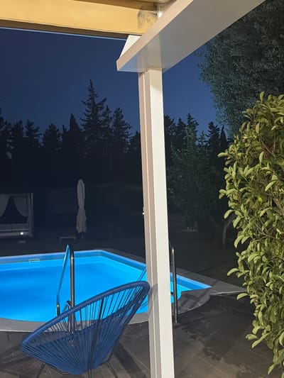 The image shows a nighttime view of an outdoor swimming pool and patio area. A bright blue swimming pool with underwater lighting is partially visible in the foreground and midground. A blue Acapulco-style chair is positioned in front of the pool. A white support beam of a modern awning or pergola stands in the center of the image, obscuring part of the view. Beyond the pool, a canopy bed with white curtains and a folded white umbrella are visible. The background consists of dark silhouettes of pine trees against a deep blue night sky. Lush green foliage from a large plant or tree is prominent on the right side of the image. The scene appears to be a private residence or resort in Lloseta, Spain, characterized by its tranquil nocturnal ambiance. There are no people or animals present in the frame.