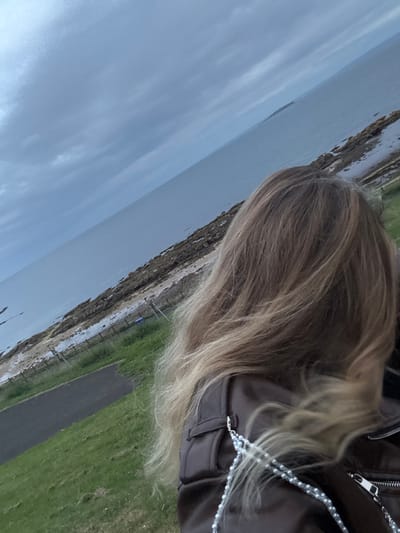 A scene captured from a rotated angle shows a woman with light brown and blonde hair wearing a brown leather jacket. The background depicts a coastal landscape with a cloudy sky and a body of water on the left. A rocky shoreline transitions into a grassy area on the right, alongside a path. A thin, delicate string with beads is visible on the jacket.