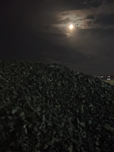 The image depicts a dark, nocturnal outdoor scene, oriented vertically with a pile of coarse, dark aggregate dominating the right side of the frame. This material, resembling crushed stone, gravel, or possibly coal, forms a textured mound with individual pieces reflecting minimal light.
In the upper-left portion of the image, a bright, circular light source, presumably the moon, shines through a haze of clouds, casting a soft glow that illuminates portions of the sky. The clouds appear streaky and somewhat ethereal. Below the moon, a faint, vertical, indistinct shape can be discerned, possibly representing a person standing or walking, blurred by low light or movement.
The overall setting suggests an industrial area, a construction site, or a railway yard, particularly given the large quantity of aggregate material. The time of day is clearly night, characterized by deep shadows and limited visibility. The atmosphere appears hazy or misty, likely due to cloud cover around the moon. In the far top-right corner, a few small, bright points of light are visible, hinting at distant structures or machinery. There is no visible text in the image. The scene conveys a somewhat desolate and mysterious mood, typical of an industrial environment after dark.