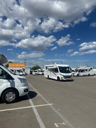 The image depicts a parking lot filled with recreational vehicles (RVs) in Quart de Poblet, Spain. The RVs are predominantly white. An orange billboard with "ac-llar.com" is visible in the background. The sky is blue with puffy white clouds. Power lines stretch across the horizon. The parking lot is marked with white lines, and the parking space numbers are visible. The image appears to have been taken on a sunny day.