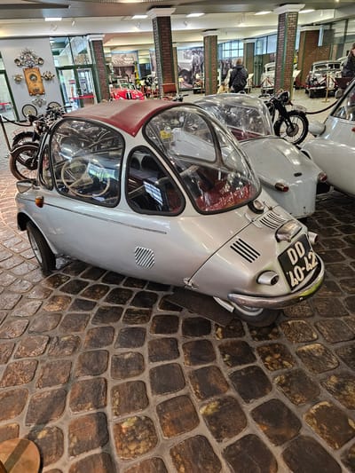 The image shows a light-colored vintage car, likely an Isetta or a similar microcar, parked on a cobblestone floor in what appears to be a museum or exhibition space. The license plate on the car reads "DD 40-42". Adjacent to it are other vintage vehicles and motorcycles. In the background, architectural details such as pillars, windows, and possibly a hallway can be seen. The city of Архангельское, Russia is not explicitly visible in the image, but it can be inferred based on metadata, if any. The overall lighting suggests an indoor setting with controlled lighting, possibly a museum display.