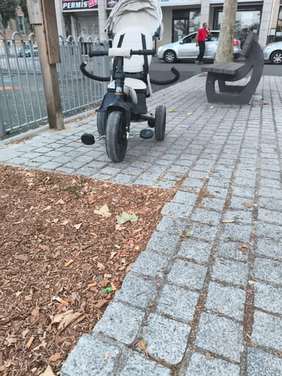 The image depicts a street scene in Carrières-sous-Poissy, France. In the foreground, a child's tricycle with a white canopy is positioned on a sidewalk paved with grey cobblestones. The ground beside the sidewalk is covered with a layer of dry brown leaves and debris, with several discarded cigarette butts visible among them. To the right, a dark grey, modern-style bench is situated on the cobblestone pavement. In the background, a silver car is parked, and a man in a red shirt and dark pants is walking away from it. A gray metal fence runs along the left side of the image, behind which is a street with some parked vehicles. Buildings are visible further in the background. The scene appears to be outdoors during daylight hours.