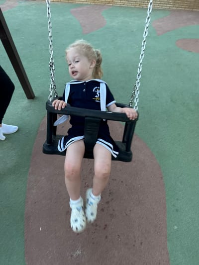 A young child with blonde hair pulled into a ponytail is swinging on a dark-colored swing. The child is wearing a navy blue sailor-style dress with white trim and white socks with white clogs. The child's knees are visible and appear to have minor abrasions. The swing is suspended by metal chains from a dark brown swing set frame. The ground beneath the swing is a rubberized surface with patches of green and brown. In the background, a brick wall is visible. To the left of the swing set, a person's legs and feet are visible, wearing black pants and white sneakers. The scene appears to be a playground.
This image was taken in the city of Sant Feliu De Guíxols, Spain.