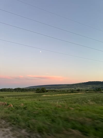 The image depicts a landscape scene with power lines stretching across the sky. The sky transitions from a pale blue near the horizon to a lighter shade at the top, with wisps of pink and orange clouds suggesting a sunset or sunrise. A faint moon is visible in the upper left quadrant of the sky. To the right of the sky is a sloping field of green grass, with a darker line of trees at the base of a hill. The field appears to be agricultural, with some sections showing different patterns of cultivation or growth. The overall impression is one of a rural setting, possibly in the early morning or late evening given the sky colors and the presence of the moon. The image is oriented vertically, with the sky occupying the left two-thirds of the frame and the landscape the right third. There is no visible text.