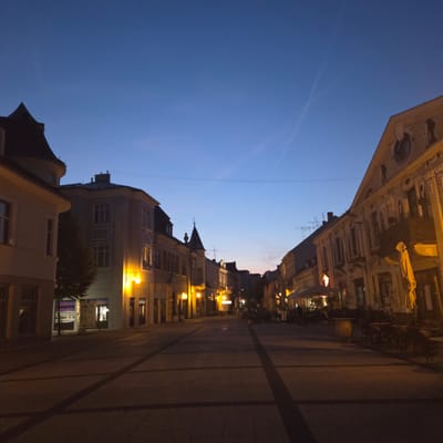 The image depicts a street scene in the city of Piešťany, Slovakia, at dusk or early night. The sky is a deep, clear blue with faint contrails visible. The street is paved with rectangular stones, with dark lines indicating a pattern. On both sides of the street, buildings with illuminated windows and decorative facades line the walkway. Some buildings feature traditional architecture with ornate detailing and pitched roofs. Cafe seating with umbrellas and chairs is visible on the right side of the street, suggesting outdoor dining areas. The overall atmosphere is quiet and serene, illuminated by streetlights and lights from within the buildings.
