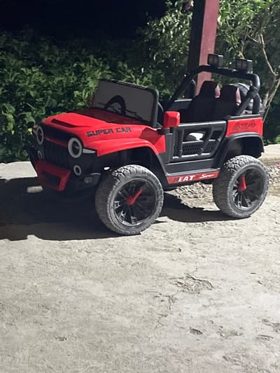 The image features a red and black toy off-road vehicle, resembling a jeep, positioned outdoors. The photograph is oriented sideways, making the vehicle appear to be lying on its right side, but it is designed to stand on four large, knobby tires with distinct red hubs.
Visible text on the vehicle includes "4X4" in white lettering on a red panel at the rear fender. On the side, above the front wheel, the word "Super" is clearly legible, with "HEAT" partially visible before it. Further forward, on the side panel, the phrase "SUPER CAR" is written vertically. The front of the toy vehicle has circular details that suggest headlights.
The setting is an outdoor environment, likely during the day, with soft shadows indicating either overcast conditions or late afternoon light. To the right of the vehicle, there is a light-colored, textured concrete or paved surface, suggesting a ground or pathway. On the left side and extending into the background, lush green foliage, likely bushes or trees, provides a natural backdrop. A dark red or brown structural element, possibly a beam or post, is partially visible on the far left. The scene is static, with no activity or people present. The location context is Jrarbi, Armenia.