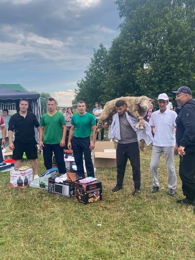 A group of men, some of whom are wearing sportswear, stand on a grassy field in Shemorbash, Russia, with a cloudy sky above and trees in the background. In the foreground, several boxes of electronics and appliances are displayed, including a microwave and what appears to be a food processor. One man in the center is carrying a sheep over his shoulders, draped in a patterned cloth. Another man, wearing a white cap, is speaking to a man in a black hoodie. The men are positioned in front of a tent structure, suggesting an outdoor event or competition.