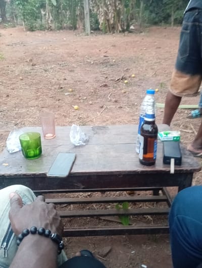 The image captures a casual outdoor gathering, focusing on a rustic wooden table laden with items, seen from a low perspective. The setting appears to be a dirt or earthy ground, common in rural or natural environments, with numerous small, spherical yellow objects (possibly fallen fruits like lemons or small oranges) scattered across it. In the background, lush green vegetation, including what look like banana trees and other dense foliage, suggests a tropical or semi-tropical climate. The lighting indicates it's daytime, possibly late afternoon or early morning, with soft, diffused light and no harsh shadows.

On the dark wooden table, which shows signs of wear, several objects are arranged. From left to right, there is a green translucent glass tumbler and a pink or amber transparent glass. Next to these, a dark-colored smartphone lies face up on the table. A crumpled silver/white plastic bag or wrapper is also visible. Towards the right side of the table, a blue plastic water bottle stands next to an amber glass bottle, likely containing beer, with a partially visible label. Further to the right, a small green rectangular box (possibly cigarettes or matches) rests beside a black power bank or portable charger. A small white object, resembling a rolled cigarette or cigarette butt, is seen protruding from the side of the power bank.

Parts of at least two, possibly three, individuals are visible, suggesting a social interaction. In the immediate foreground on the left, a dark-skinned hand and forearm are seen resting on a leg. The person is wearing light-colored trousers or jeans with a distinct zipper detail on the side. A black beaded bracelet is worn on the wrist. To the right, slightly behind the table, the lower half of another person is visible, standing or sitting. This individual has dark skin and is wearing blue denim shorts that are faded or bleached to a light brown/tan color at the bottom. Their feet, wearing sandals or flip-flops, are also visible. A third person is hinted at by a portion of a leg in blue trousers visible in the bottom right corner of the frame.

The overall scene conveys a relaxed atmosphere, likely a casual hangout among friends, enjoying drinks and conversation in an unpretentious outdoor setting.