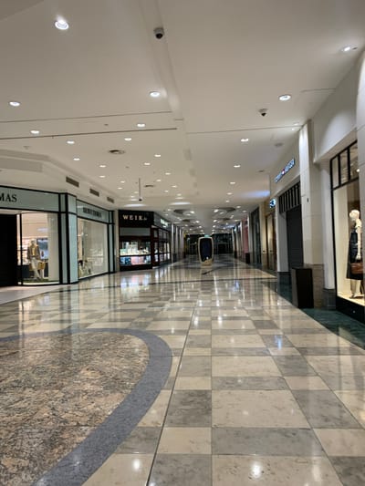 This is an indoor view of a shopping mall hallway. The hallway has a white ceiling with evenly spaced recessed lights. Security cameras are also visible along the ceiling. Shopfronts line both sides of the hallway, including "WEIR.ie" and "BROWN THOMAS." The floor is tiled in a checkered pattern of alternating light and dark squares, with a decorative circular pattern in the center. At the end of the hallway, a single vehicle is visible. Based on the visible retailers, this photo appears to be taken inside Dundrum Town Centre, located in Dundrum, Dublin 16, Ireland. No people are visible in the photo.