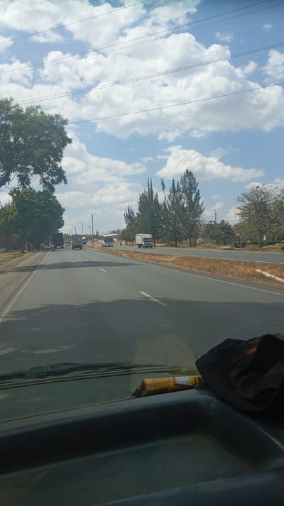 A road stretches into the distance under a blue sky with scattered clouds. Several vehicles, including trucks and a bus, are visible on the road, traveling in both directions. Trees line the side of the road. The view is from inside a vehicle, with the dashboard visible in the foreground, including a tube and some cloth. Power lines stretch across the sky.