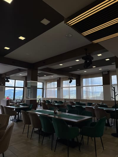The image shows the interior of a restaurant in Sapareva Banya, Bulgaria. Rows of tables with green runners and chairs are visible. Large windows reveal a mountain view outside. The ceiling has a dark brown and white geometric design with recessed lighting. A black fixture is attached to the ceiling.
