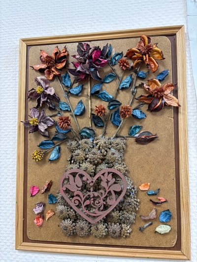 An upright, rectangular artwork crafted from dried natural materials, encased in a light-colored wooden frame, is depicted against a textured white wall in Novosergievka, Russia. The artwork's background is a plain, light brown board. The central subject is a vase-like form constructed from numerous small, clustered, light brown dried seed pods or florets. Superimposed on the upper portion of the vase is a pink, decorative, heart-shaped cutout featuring an ornate scroll pattern. Extending upwards from the vase are multiple thin, bare stems, each adorned with various dried botanical elements. These include several prominent dried flowers in shades of faded orange, deep magenta, and pale purple, some with distinct yellow centers. Interspersed among the flowers are numerous small, oval-shaped dried leaves, coated with a metallic blue-green finish. Additionally, smaller, spiky, reddish-brown dried flower clusters are present. At the bottom of the frame, several loose, scattered dried petals or leaf fragments in varying shades of pink, orange, blue, and cream are visible. No individuals or active subjects are present.