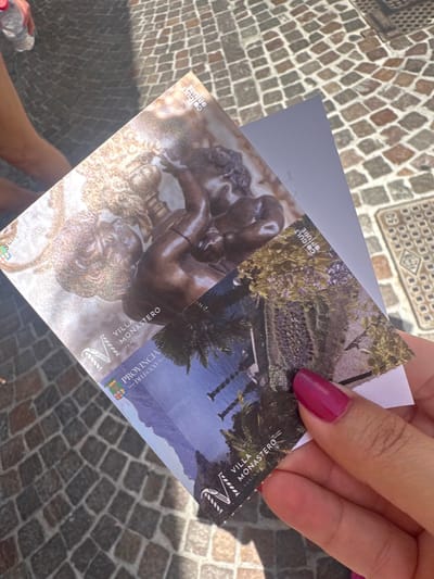The image shows a person holding two cards in front of a cobblestone ground. The card in the foreground has a picture of a lake and trees and says "VILLA MONASTERO" and "PROVINCIA -DI LECCO". The card behind it has a picture of a sculpture. The cobblestone ground appears to be in an outdoor setting. The person's hand holding the cards has a bright pink fingernail polish. Based on the text on the card, the location is Villa Monastero in Varenna, Italy, which is part of the Province of Lecco.