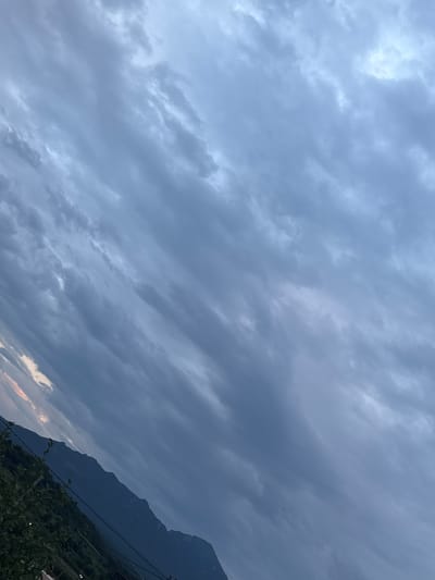 The image is a slightly tilted, full outdoor shot of a mountainous landscape under a cloudy sky. In the foreground, on the left side of the image, the dark green foliage of trees and shrubs is visible, with some thin, dark wires also present. Behind the vegetation, the dark blue silhouette of a mountain range stretches across the frame, with its peaks and slopes clearly defined against the sky. The sky itself is filled with a multitude of gray and blue clouds, with some lighter areas suggesting breaks in the cloud cover or the glow of diffused light. The overall impression is one of a dramatic, overcast sky over a rugged terrain. The location is identified as Novoteretsky, Russia. There are no people or specific actions depicted in the image.