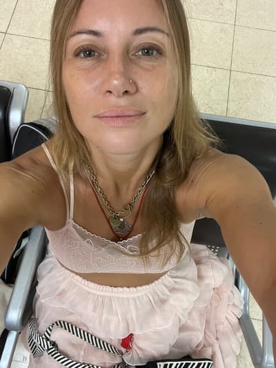 A woman with blonde hair and green eyes is pictured from the chest up. She is wearing a light pink, ruffled dress with lace detailing and a gold necklace. She has a small gold hoop in her nose. She is sitting in what appears to be an airport waiting area, indicated by the tiled floor and dark seating. The image is a selfie, with the woman looking directly at the camera. The lighting suggests it could be daytime. The overall mood seems relaxed, perhaps indicating travel. There is no visible text in the image.