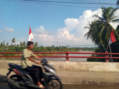 In Kulukuta, Indonesia, a man rides a motorcycle across a bridge. The motorcycle is in motion, creating a blur of movement. The man is wearing a light green shirt and dark pants. In the background, a red railing lines the bridge, separating it from a river below. The river flows through a landscape of lush green trees and palm trees. The sky above is blue with scattered white clouds. Two Indonesian flags, red and white, are visible, one on a flagpole to the left of the frame and another attached to a palm tree on the right. A transmission tower can be seen in the distance. The road surface of the bridge is made of concrete and dirt.
