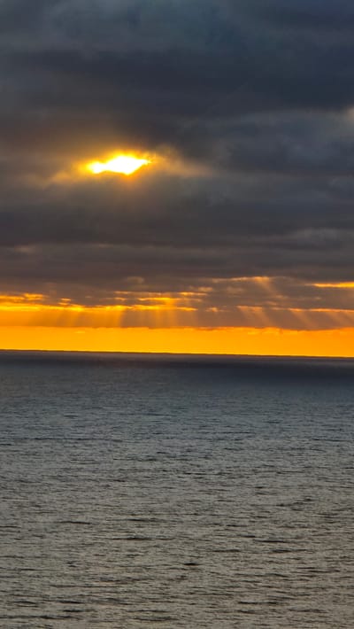 The image shows a dramatic sunset over the Pacific Ocean. Dark, heavy clouds dominate the upper portion of the sky, with a bright, golden light breaking through a gap in the clouds, casting rays downwards. Below the clouds, a vibrant band of orange and yellow stretches across the horizon, illuminated by the setting sun. The ocean is a deep blue-grey, with a rippled surface reflecting the colors of the sky. The scene appears to be captured from a high vantage point, possibly a cliff or bluff overlooking the coastline in Encinitas, California. There are no people, subjects, or objects visible in the frame, only the natural elements of sky and sea.