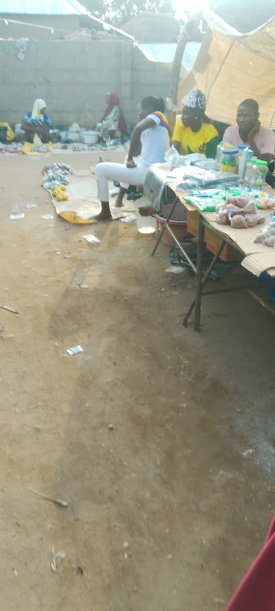 This image captures a scene at an outdoor market in Doka-Tofa, Nigeria. Several people are present, including women in traditional headscarves and men. One woman in a blue and yellow patterned garment is seated, possibly selling goods. Another woman in a maroon headscarf is also seated. A young man wearing a white t-shirt and white pants is sitting on the ground, looking towards a table laden with items. Nearby, two other individuals are seated at the table. One is wearing a bright yellow t-shirt and the other a pink shirt.
The market appears to be set up on a dirt ground, with various items displayed on a table and scattered around. The table is covered with what seems to be packaged goods, some in plastic wrap, and possibly food items like meat or fish. Several plastic containers and bottles are also visible on the table. The setting suggests a bustling marketplace, likely in the morning or late afternoon, given the natural lighting. The presence of a makeshift shelter with a tarp in the background hints at the temporary nature of some stalls. The overall atmosphere appears casual and transactional, characteristic of a local market day. No discernible text is visible in the image.