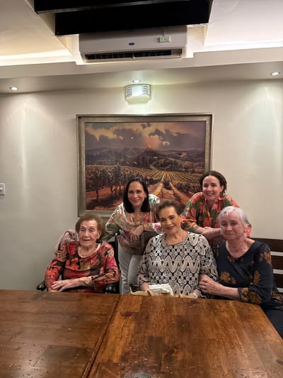 Five women are posing for a photograph at a large, wooden table in what appears to be a dining room or restaurant. Behind them is a painting of a vineyard landscape. Above the painting is a light fixture, and above that, an air conditioning unit mounted on the ceiling. The location is presumed to be in Tijuana, Mexico based on the information provided. The women are wearing various patterned tops, and some are seated while others are standing or leaning against the table.
