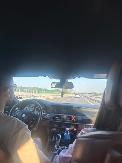 The image shows the interior of a car being driven on a highway, likely in the vicinity of Trstenik Municipality, Serbia, based on the context provided. The perspective is from the rear passenger seat, looking forward. A man with glasses is visible in the driver's seat, wearing a light-colored t-shirt and shorts. A woman wearing a pink hooded sweatshirt is seated in the front passenger seat. The car's dashboard displays various controls, including the steering wheel with a BMW logo, a speedometer, and a central infotainment screen. A water bottle is placed in a cup holder on the center console. Outside the car, a highway with multiple lanes is visible, bordered by a safety barrier and greenery. The sky is clear and blue. The overall scene depicts a journey in progress on a sunny day.