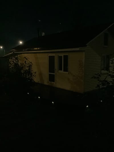 The image depicts a nighttime scene of a rural or suburban dwelling in Nikul'skoye, Russia. The structure appears to be a single-story house with a dark, sloped roof. The exterior walls are light-colored, possibly painted wood or vinyl siding, and show horizontal siding details. There are several windows visible; one large window with a dark pane is on the right side of the house, and a narrower window with a white frame and dark pane is on the left. A glass-paneled door, also with a dark pane, is located next to the window on the left. Foliage, including a small tree or bush with dark leaves, is present in the foreground and partially obscures the lower portion of the house. Several small, illuminated garden lights are scattered in the dark foreground, casting a subtle glow. The background is dark, with faint indications of distant lights and a streetlamp. The overall impression is of a quiet, dimly lit residential area at night. There are no people or animals visible in the image.