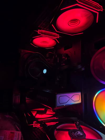 This is a photograph of the inside of a computer tower, illuminated by vibrant red and rainbow LED lighting. The central focus is a graphics card with the word "INFINITY" visible on its shroud. Surrounding it are several computer fans, some with striking red lighting and angular designs, while others are illuminated with a spectrum of colors. A CPU cooler with a fan and tubing is also visible. The overall scene is dark, highlighting the glowing components within the computer. The computer parts suggest a gaming or high-performance PC. The lighting creates a dramatic and futuristic aesthetic. The visible text indicates that this is a computer component, potentially a graphics card, with some manufacturing or model information. The location context of Aleksandrovo, Bulgaria, does not provide specific visual cues within the image itself.