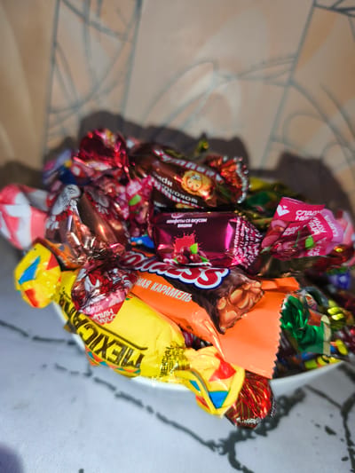 The image shows a white bowl filled with various wrapped candies. The candies are in different wrappers, including red, gold, pink, yellow, and orange. Some wrappers have text and images on them. One candy wrapper is yellow and red with the word "MEXICO" visible. Another wrapper is pink and features an image of cherries and the text "КОНФЕТЫ СО ВКУСОМ ВИШНИ" (candies with cherry flavor). The bowl is placed on a white surface with a textured pattern. In the background, a wall with a patterned wallpaper and shadows is visible. The lighting is dim, and the image is slightly out of focus. There are no people or animals present in the scene. The location of Rahachow, Belarus, is not depicted or suggested in this image.