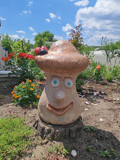 In Krutinskiy, Russia, a whimsical mushroom-shaped garden sculpture with a face is positioned outdoors on a sunny day. The sculpture, made of concrete or plaster, sits atop a tree stump and is adorned with two ladybugs on its cap. It has large, round, blue eyes, a prominent nose, and a wide, smiling mouth. The background features lush green foliage, including a bush of orange marigolds with a butterfly resting on it, and red roses. Beyond the garden, a clear blue sky with scattered white clouds is visible, along with hints of rural structures and utility poles. The ground around the sculpture is bare soil, with some fallen flower petals scattered on it, and small green plants growing in clusters. The overall scene suggests a well-maintained garden with a playful and decorative element.