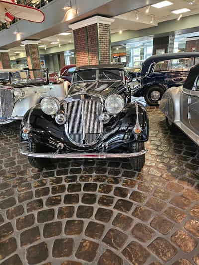 The image shows the front view of several vintage cars, with a black car prominently in the foreground, in the city of Архангельское, Russia. The cars are displayed indoors, likely in a museum or showroom, indicated by the brickwork design on pillars and ceiling structures, along with display lighting. The ground is paved with cobblestones. Other cars are partially visible behind the black one.