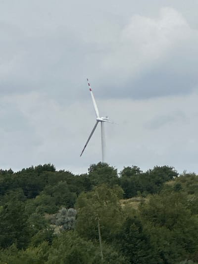 A wind turbine stands tall against a cloudy sky, with its white tower and blades clearly visible. The blades are tipped with red and white stripes, suggesting they are either painted for visibility or part of the turbine's design. The turbine is situated behind a dense line of lush green trees that cover rolling hills, indicating a rural or semi-rural environment. The overall scene is one of a single wind turbine in a natural, vegetated landscape, under an overcast sky. The image is taken from a distance, focusing on the turbine as the primary subject against the backdrop of nature. The location is specified as Kolubaivtsi, Ukraine, placing the scene within a geographical context.