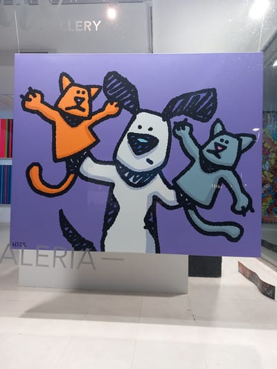 A painting of a dog with two cats is displayed in the window of Galeria Alerta in Port D'andratx, Spain. The painting features a white dog with black spots and ears, and two cats: one orange at the bottom and one grey on top. The background of the painting is purple. The name "HECK" is signed on the bottom right corner. The image is taken through the glass of the gallery's window.