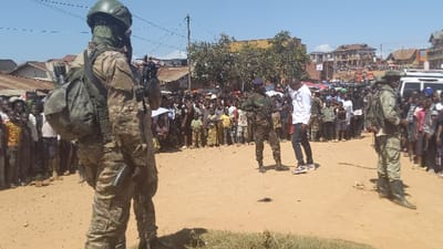 A crowd gathers in a dusty street, observed by soldiers in camouflage gear. One soldier is prominently in the foreground, facing away from the camera, with a helmet and backpack. Another soldier stands amongst the crowd. A man in a white shirt and black pants appears to be addressing the crowd, with houses and shops lining the street in the background.