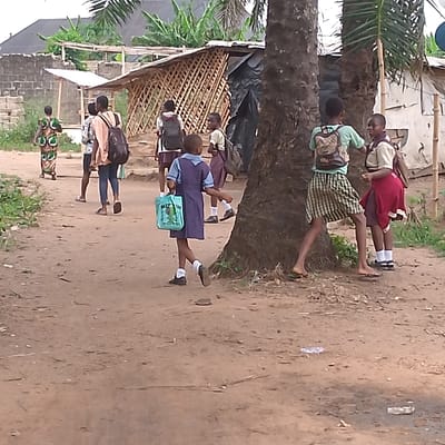The image shows a group of children walking along a dirt path in Adanisievbe, Nigeria. The children are wearing school uniforms. The girls are wearing blue dresses, and some are carrying bags. The boys are wearing khaki shorts and shirts. There is a large tree in the center of the image, and some buildings are visible in the background. The children are walking in the same direction. The group is walking away from the camera.