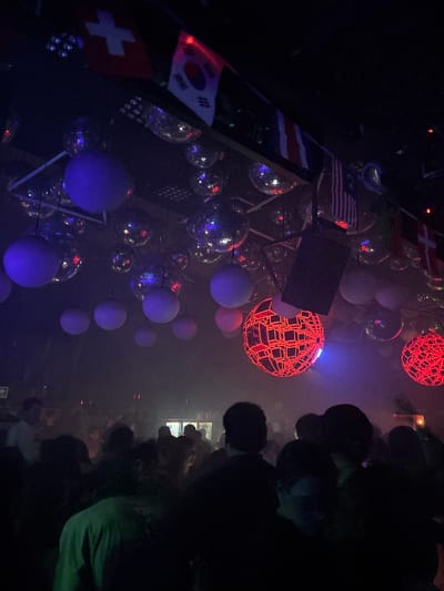 The image depicts an indoor scene, likely a nightclub or event space, with a crowd of people in the foreground. The atmosphere is dimly lit and smoky, with several large disco balls and illuminated spherical decorations hanging from the ceiling. In the background, various national flags are visible, including the Swiss, South Korean, and possibly others. Two prominent, geometric, red-lit spherical decorations are a focal point in the mid-ground. The people in the foreground are largely silhouetted, with some individuals appearing to be dancing or socializing. The presence of national flags suggests a theme of internationalism or a gathering of people from different countries. While the image strongly suggests a lively, festive environment, its specific location in the city of Veracruz, Mexico, cannot be determined from the visual evidence alone.