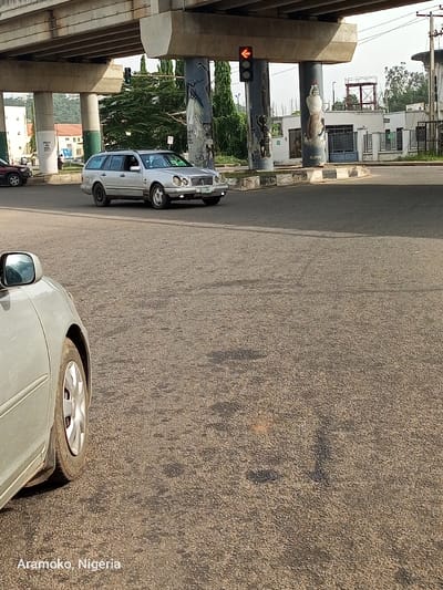 A silver Mercedes-Benz sedan is driving on an asphalt road in Ado Ekiti, Nigeria, under a bridge. The car is driving away from the camera on a road that curves to the right. The car is on the left side of the road. There is a traffic light with a red light showing, facing the camera. The bridge has a concrete underside with painted pillars. There are murals painted on the pillars. On the right side of the road, there are trees and buildings. The road is empty except for the Mercedes. The weather is clear and sunny. The scene is taken from the front, near the side of the road, looking toward the bridge. The time of day is afternoon.